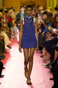 Model walks the pink runway in a cobalt blue sleeveless pinafore dress with chunky chains and a scarf, audience on both sides.