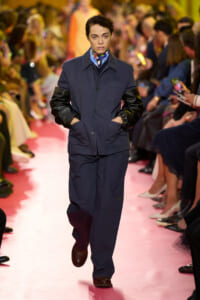 Model walks down a pink runway in a navy jacket with black leather sleeves, hands in pockets, audience watching on both sides.