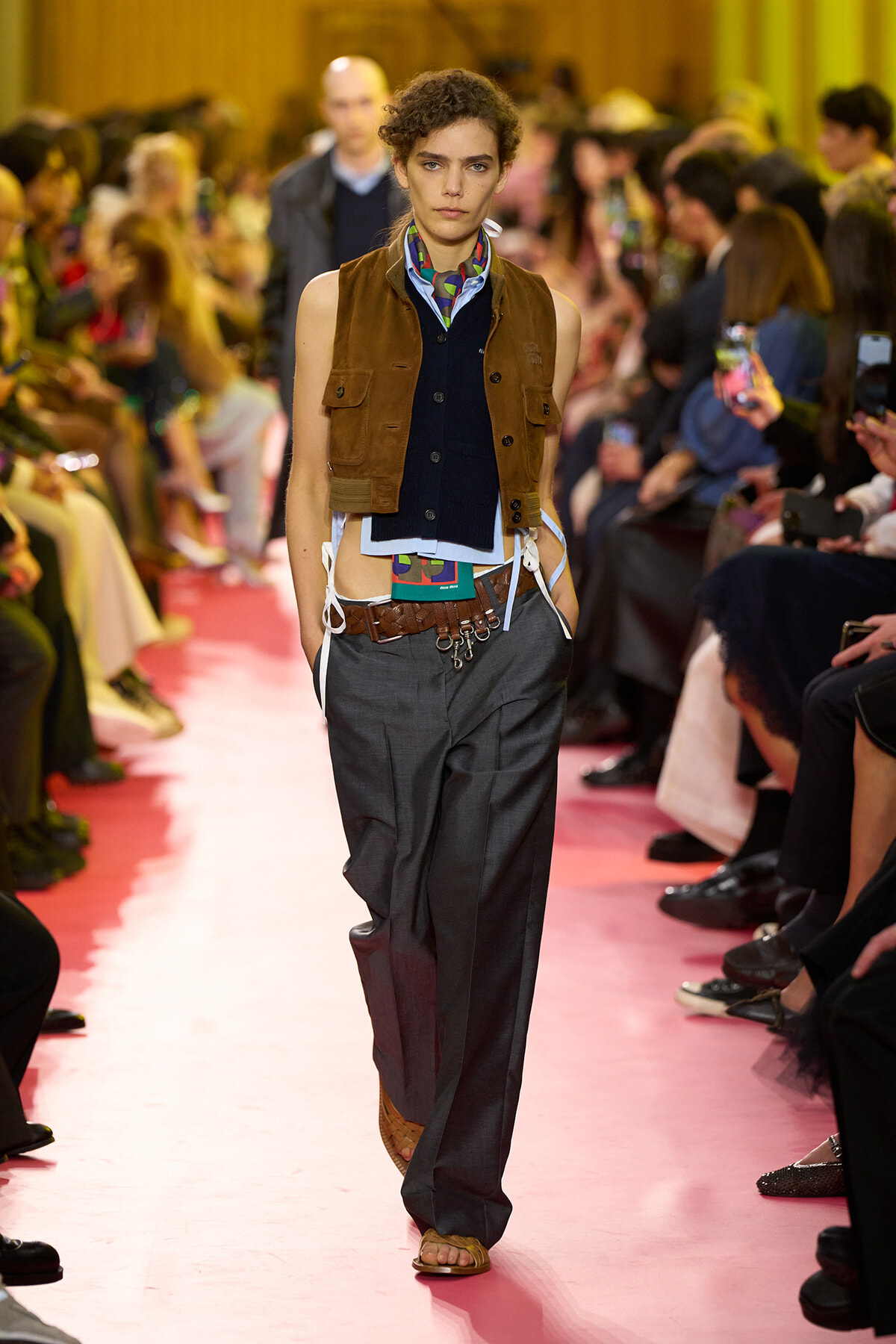 Model walks a fashion runway wearing a brown utility vest over a navy layered top, loose gray trousers, and sandals on a pink catwalk. Behind-the-scenes audience blurred.