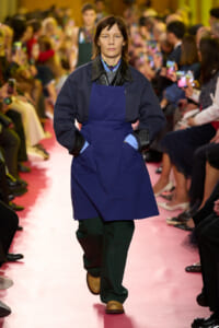Model walking down a pink runway in a blue apron over a dark jacket, with hands in pockets, audience on both sides watching and taking photos.