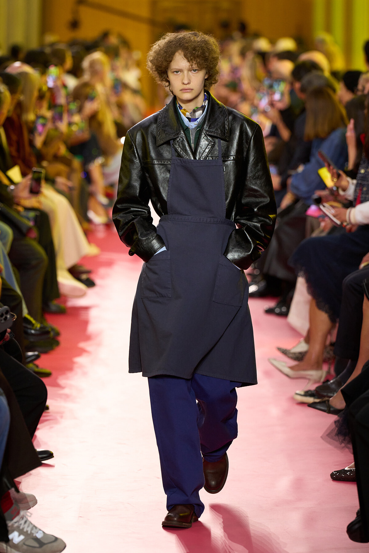 Model on a fashion-show runway wearing a navy apron over a black leather jacket, with a pink catwalk and audience in the background.
