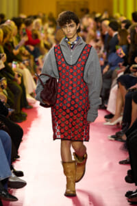 Model walks the runway in a gray zip-up hoodie layered under a red floral pinafore, carrying a burgundy bag and wearing tan boots.