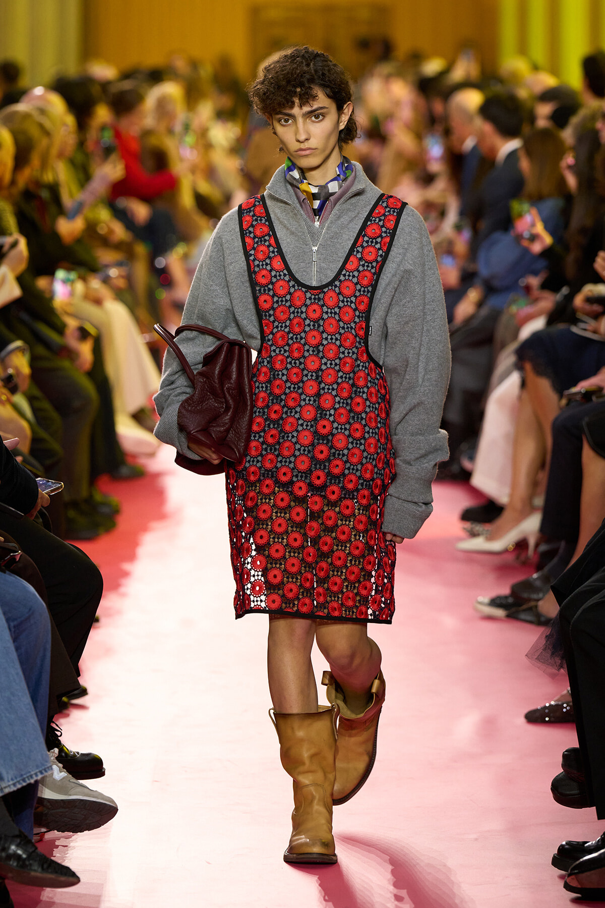 Model walks the runway in a gray zip-up hoodie layered under a red floral pinafore, carrying a burgundy bag and wearing tan boots.