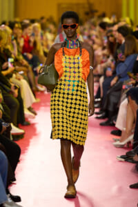 Model walking a pink runway in a bold patterned dress with orange top, circular yellow motifs, red sunglasses, and a green handbag beside her.