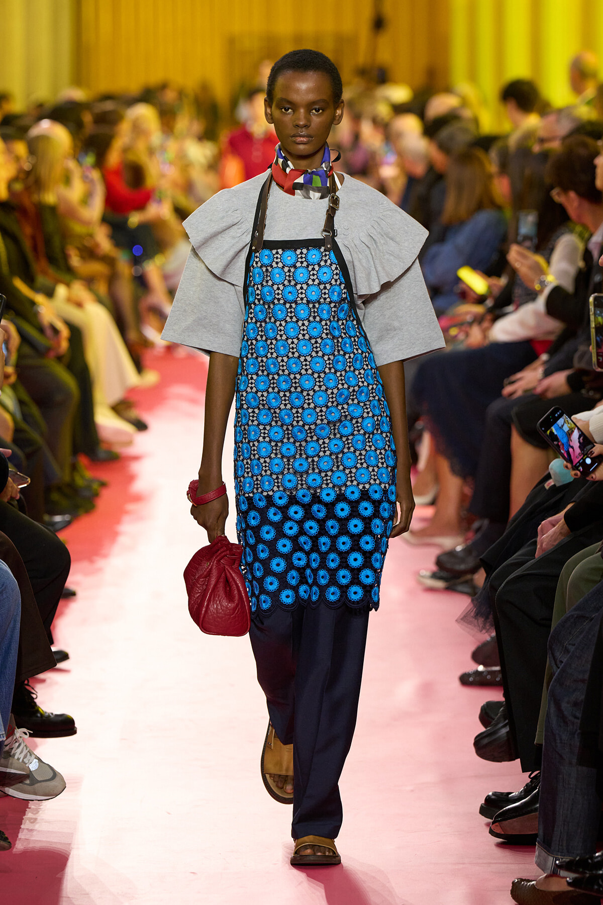 Model walking a pink runway in a blue circular-print dress over a gray cape, carrying a red handbag.