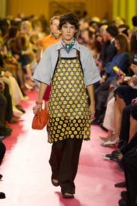 Model walks the runway in a yellow daisy-patterned apron dress over a gray top, with an orange clutch in hand.