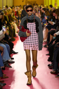 Model walks a fashion runway in a pink circular-pattern mini dress with gray sleeves, red sunglasses, and a red handbag, flanked by seated spectators.