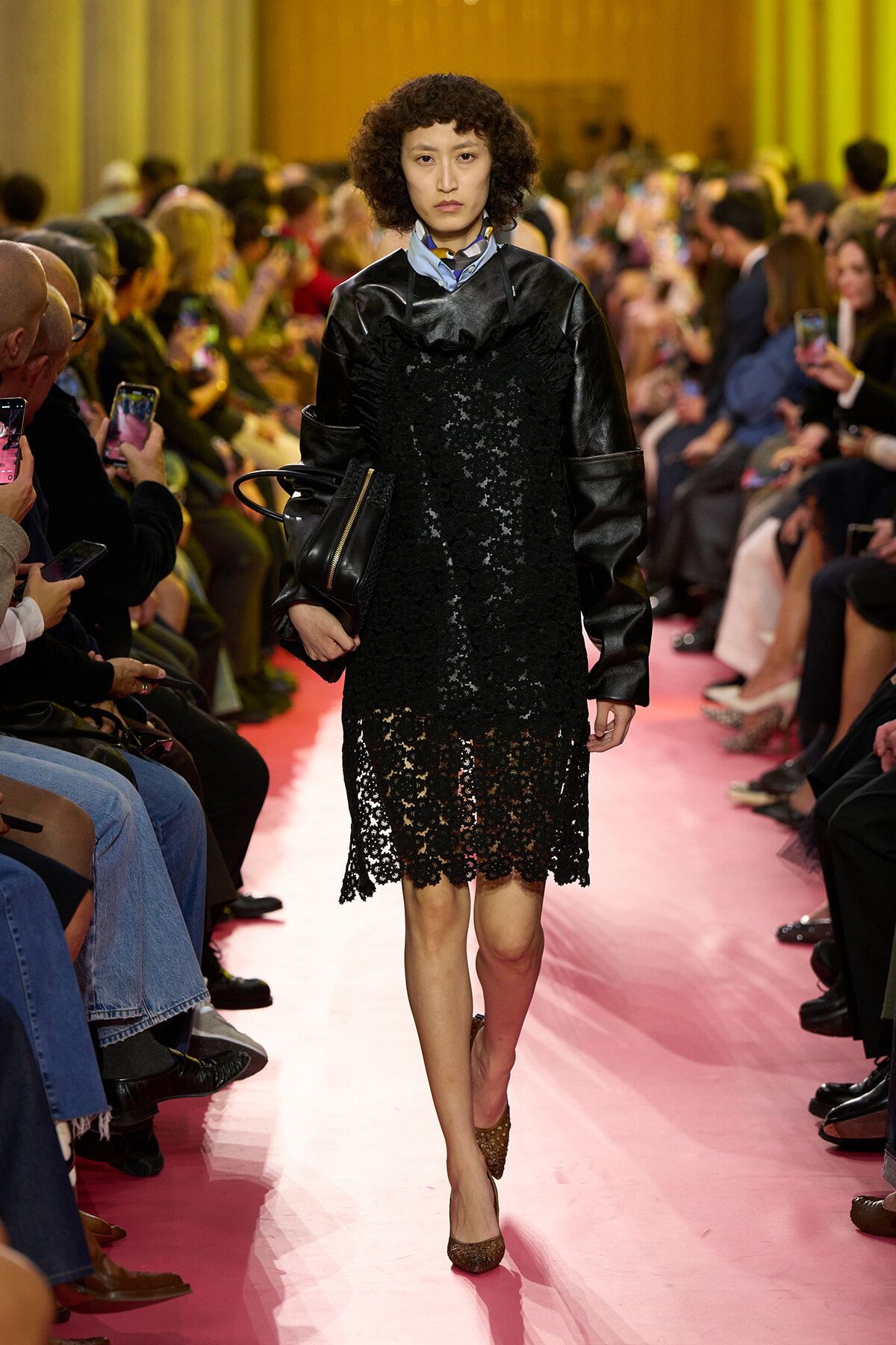 Model walks the runway in a black lace dress with leather sleeves, carrying a black handbag, on a pink catwalk with seated audience surrounding her