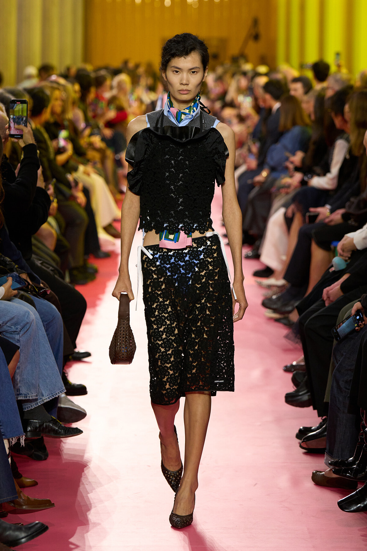 Model walks a fashion runway in a black lace two-piece outfit, pink floor, audience on both sides watching.