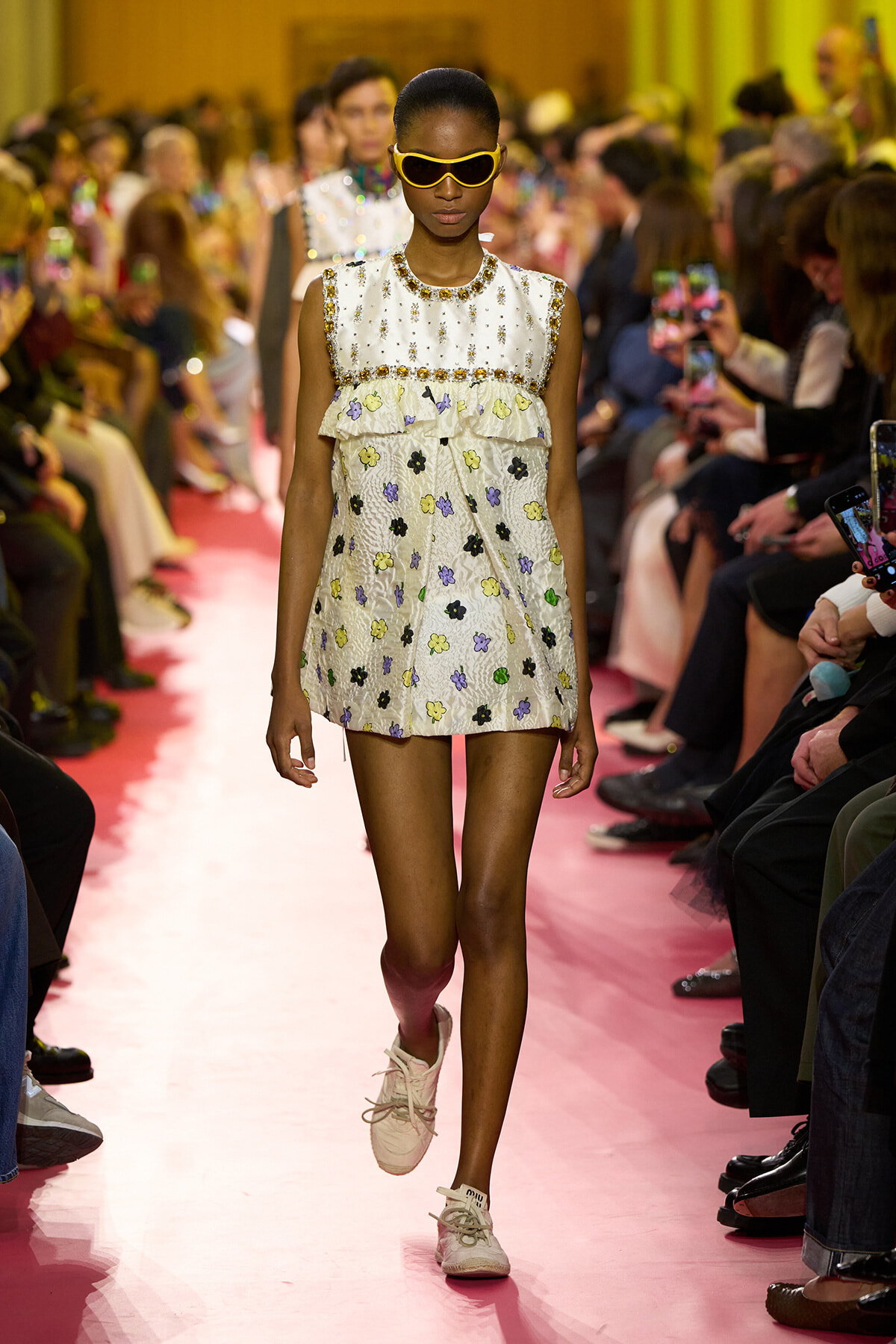 Model walking a pink runway in a white sleeveless floral mini dress with beaded yoke, yellow sunglasses, and pastel sneakers.