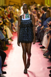 Model walks a pink runway in a navy blue lace mini dress with a black ruffle and a patterned scarf around the neck, during a fashion show.