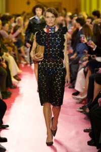 Model walks the runway in a black lace sheath dress with ruffled sleeves, a colorful scarf, and a tan clutch on a pink catwalk.