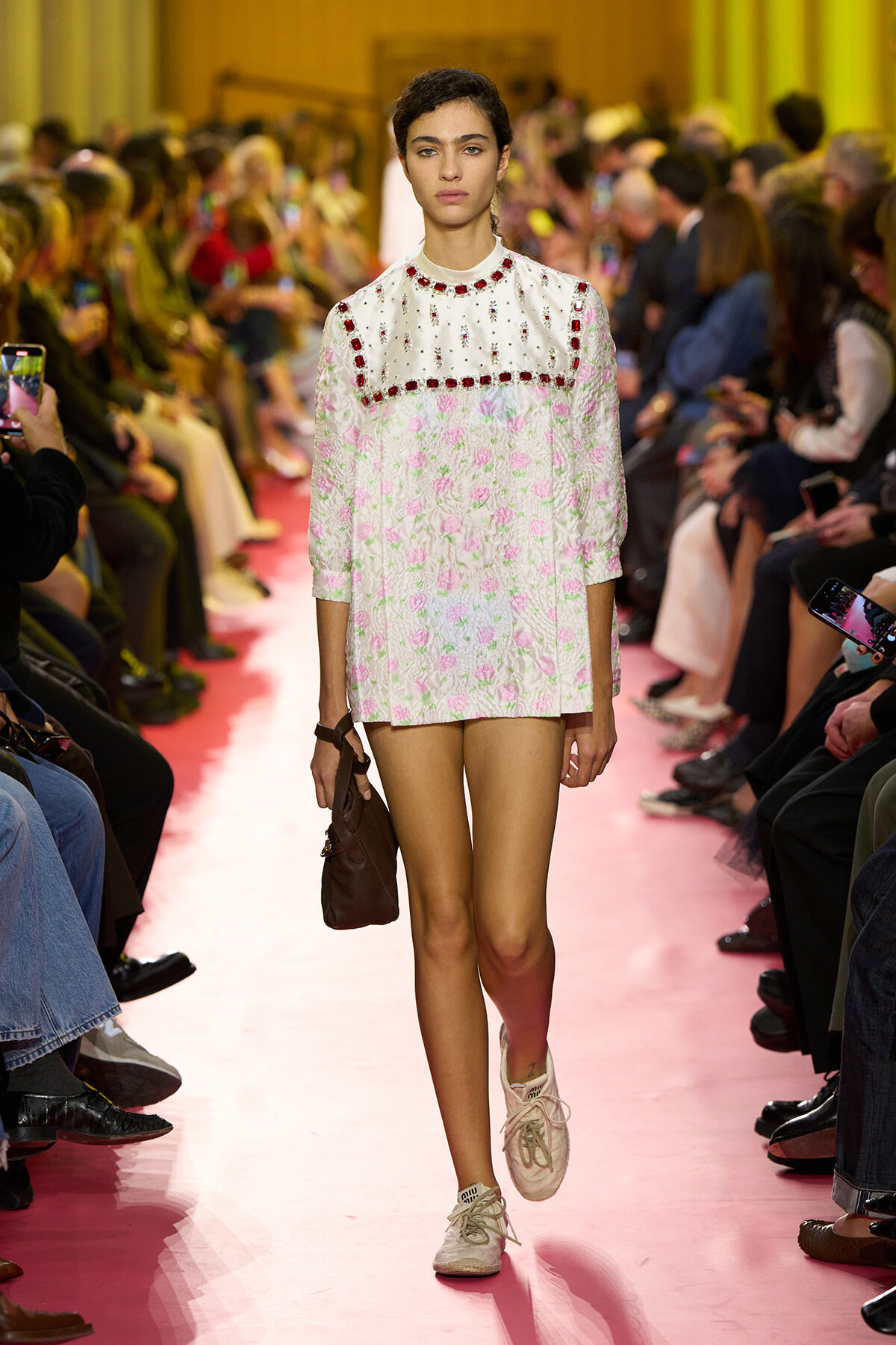 Model walks a pink runway in a white floral mini dress with red gemstone trim and a brown bag, surrounded by photographers and spectators.