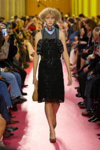 Model walking a pink runway in a black lace dress with puffed leather sleeves, a colorful neck scarf, and gold heels, carrying a woven bag.