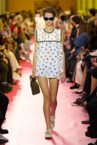 Model walking the runway in a sleeveless white dress with black bead trim, blue sunglasses, and a brown handbag.