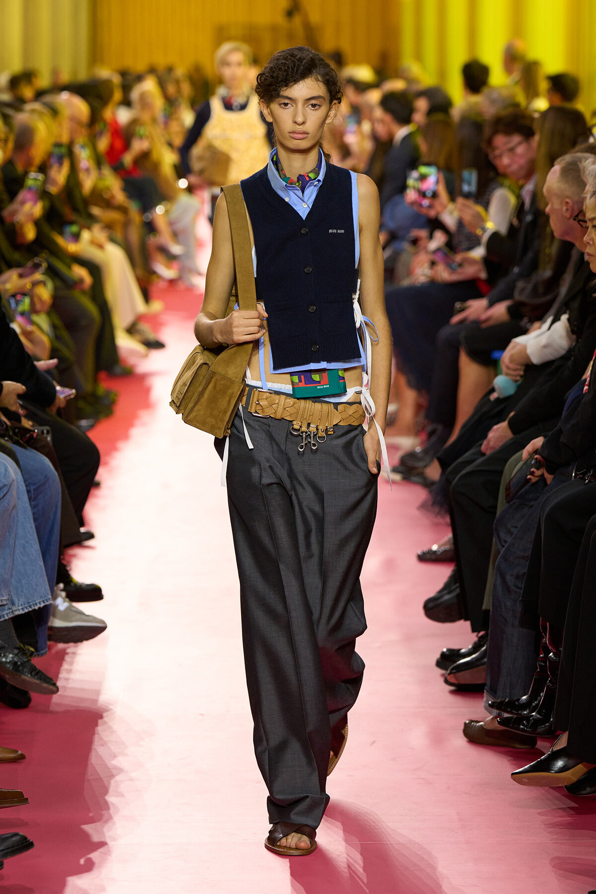 Model walking a pink runway at a fashion show, wearing a navy vest over a light blue shirt, gray wide trousers, and a tan shoulder bag.