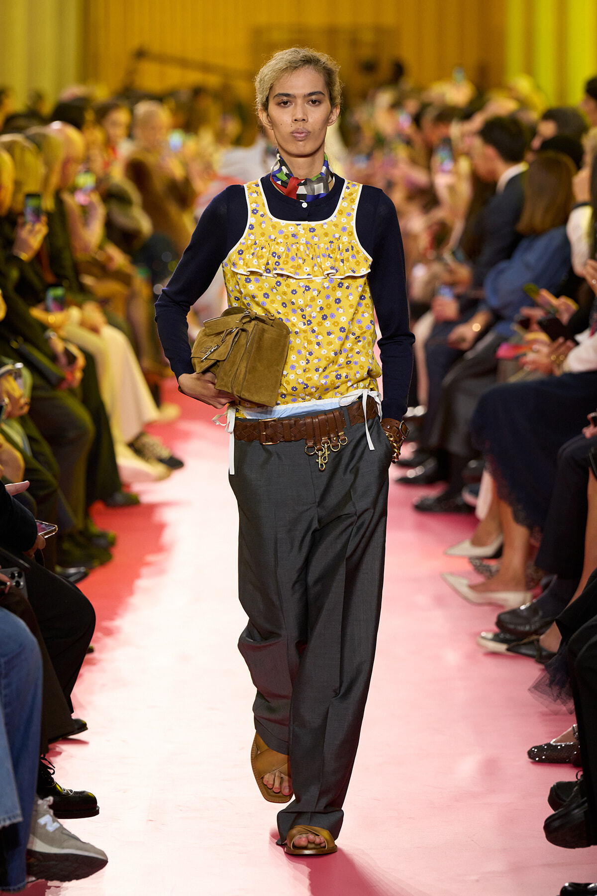Model walks the runway in a yellow floral sleeveless top over a navy long-sleeve shirt, gray baggy trousers, and a brown belt with keys, carrying a tan bag.