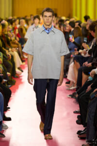 Male model walks a pink runway in a light gray short-sleeve top, blue shirt, and navy trousers, with spectators flanking the catwalk.