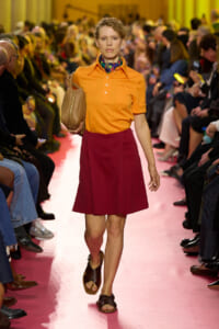 Model walks the runway wearing an orange polo tucked into a burgundy A-line skirt, carrying a tan handbag and wearing brown sandals on a pink-catwalk.