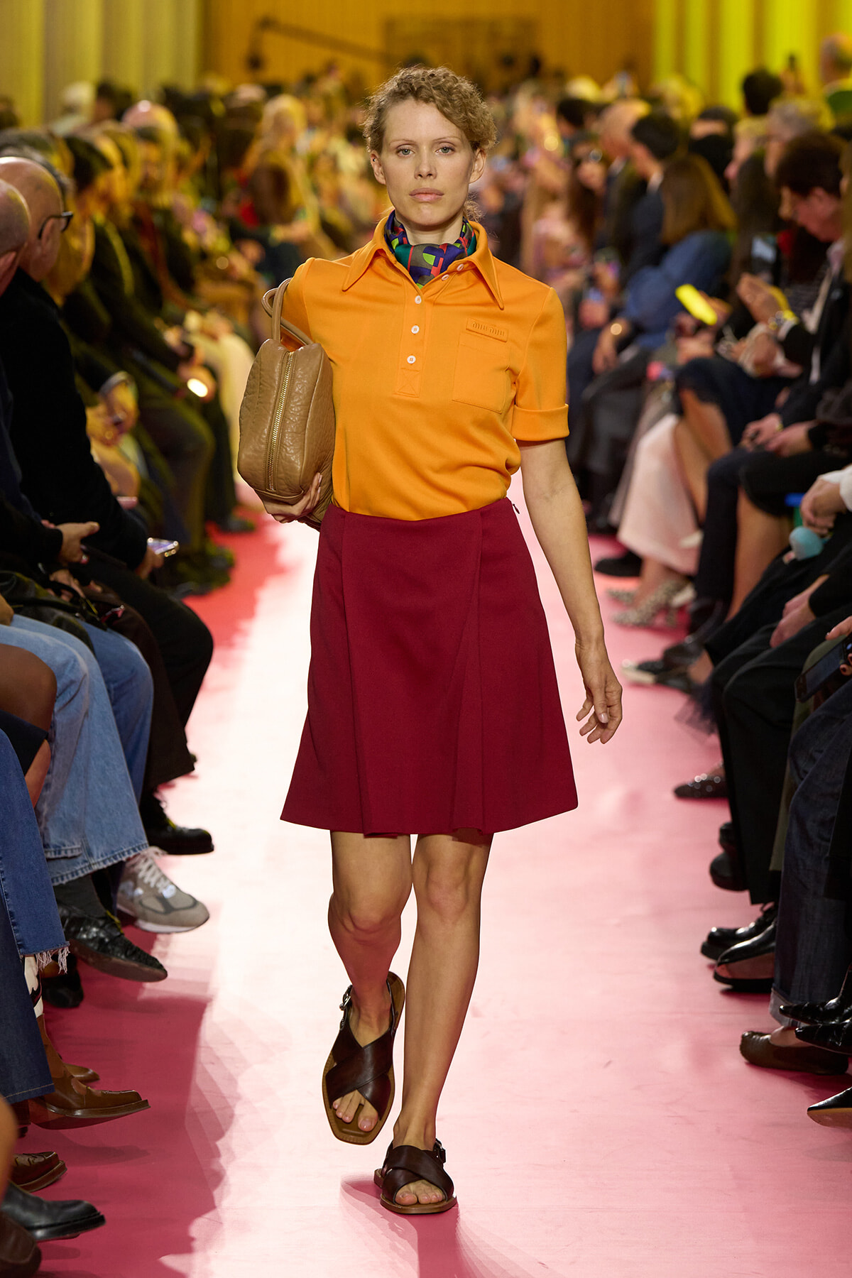 Model walks the runway wearing an orange polo tucked into a burgundy A-line skirt, carrying a tan handbag and wearing brown sandals on a pink-catwalk.