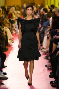 Model walks the pink runway in a black textured dress with oversized ruffle shoulders at a fashion show.