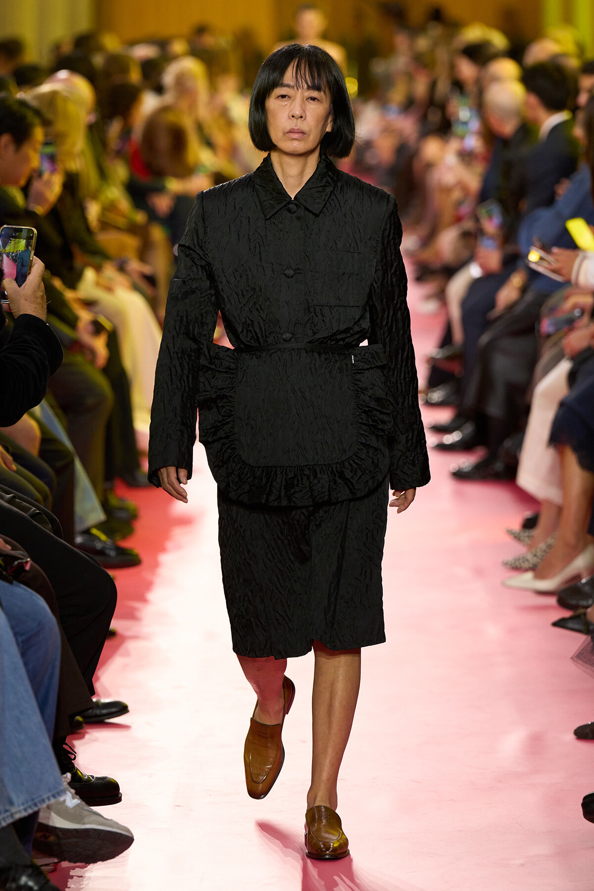 Model walks a pink runway in a black textured jacket and matching skirt, with brown loafers, as the audience watches.
