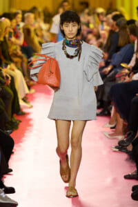 Model walking runway in a light gray oversized dress with ruffled sleeves, carrying an orange handbag and wearing bold beaded necklaces.
