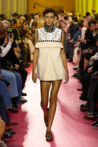 Model walking a pink runway in a cream sleeveless dress with a beaded square neckline and ruffled chest overlay.