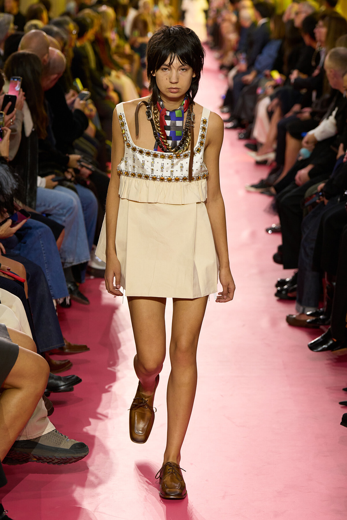 Model walks a pink runway in a cream, beaded sleeveless dress with layered necklaces and brown loafers.
