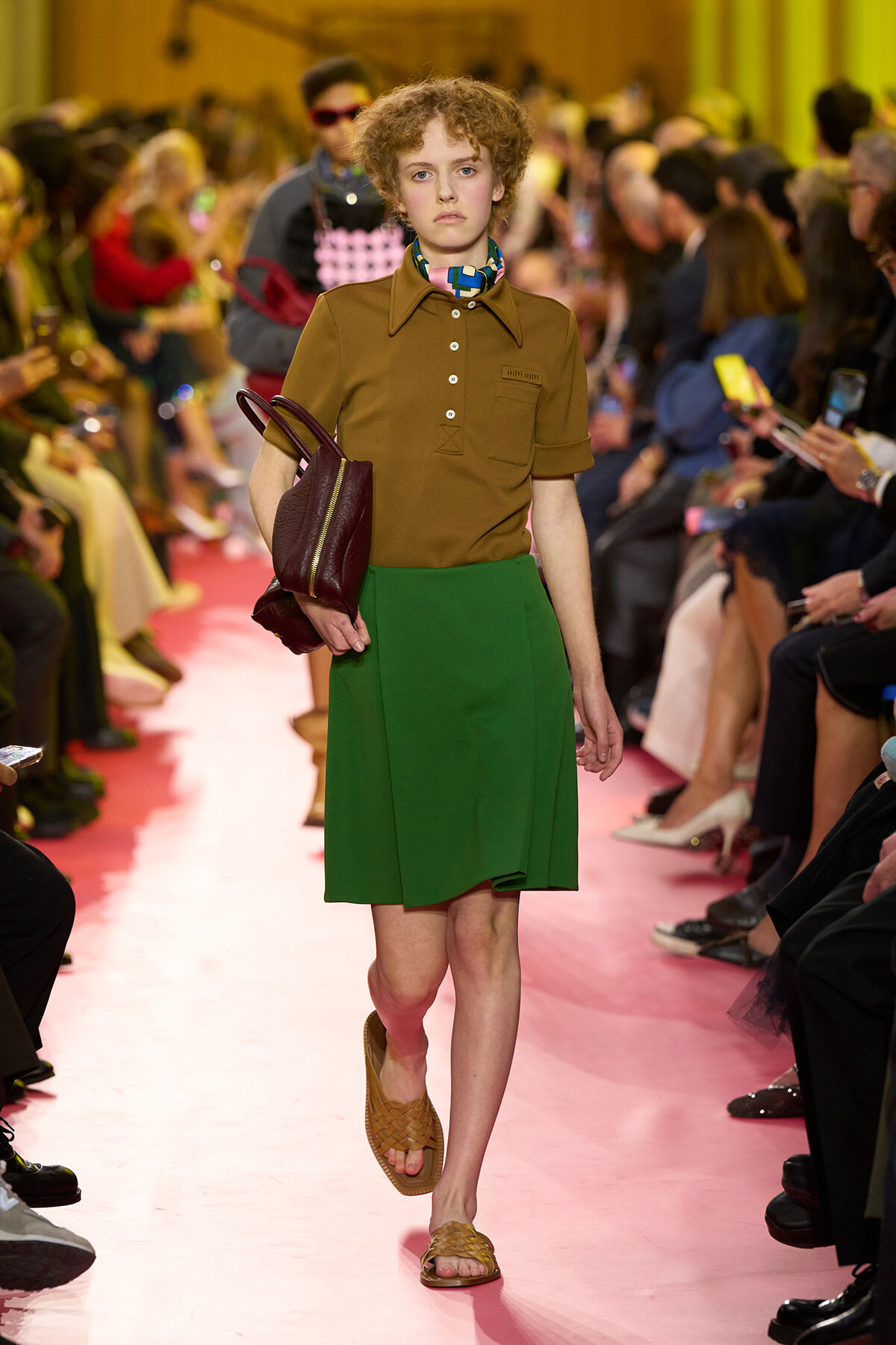 Model walks the runway in a brown short-sleeve polo, green A-line skirt, tan woven sandals, and a burgundy handbag under pink lighting.