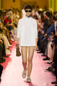 Model walking a fashion show in a white oversized tunic dress, green sunglasses, and a multicolored scarf, carrying a small brown bag and wearing beige sneakers on a pink runway.