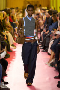 Model walks a fashion runway in a layered gray vest over a blue shirt, oversized navy pants, and a brown crossbody bag.