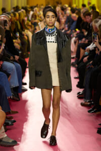 Model walks the runway during a fashion show on a pink catwalk, wearing a green oversized coat with leather details over a beige dress and a beaded collar.