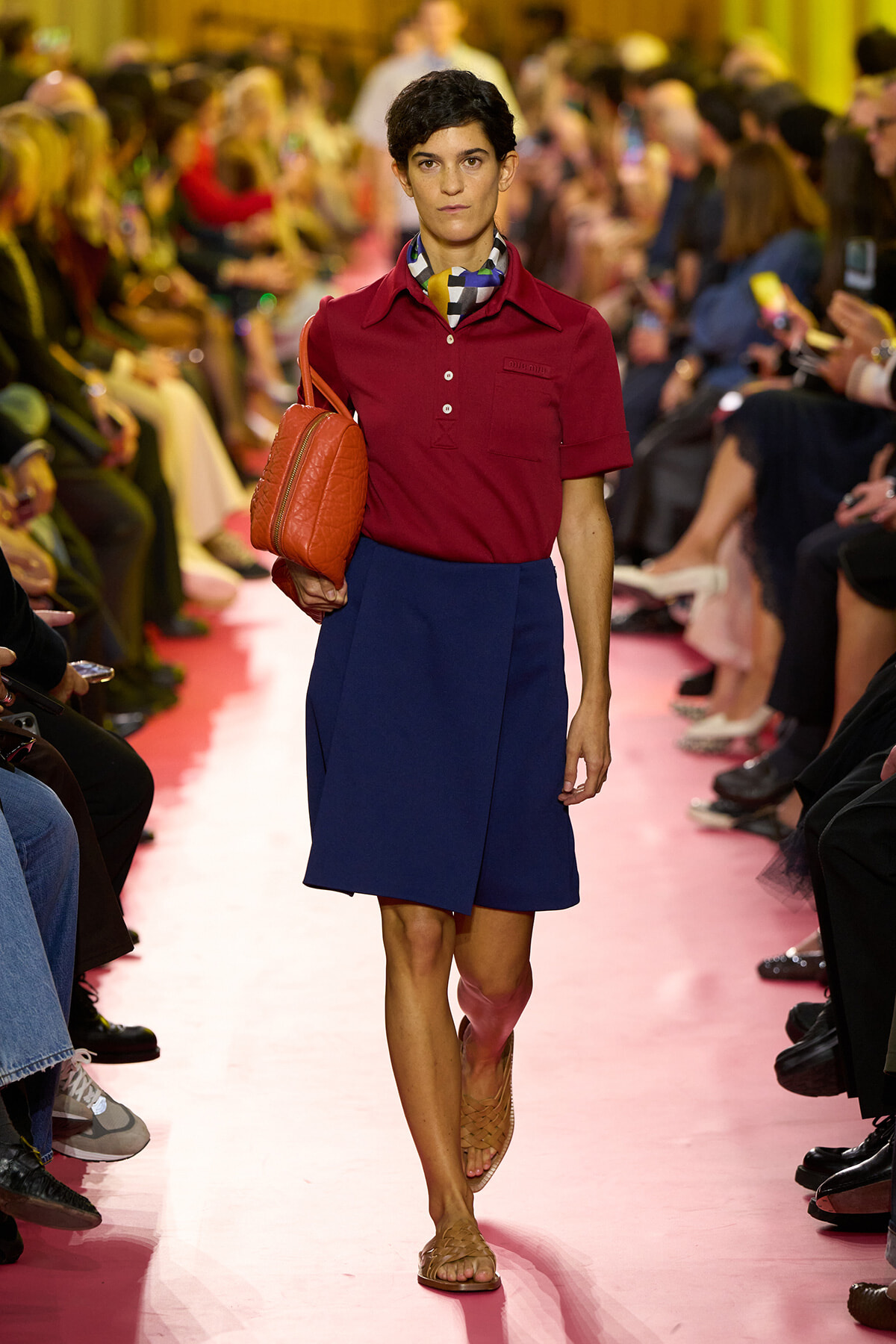 Runway model walks in a burgundy shirt, royal blue skirt, patterned scarf, and an orange handbag.