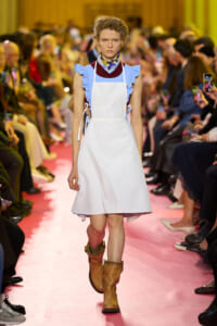Model walks a pink runway in a light blue dress with a white apron and tan boots, audience blurred in the background.