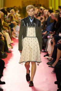 Model walking a pink runway in a dark green jacket layered over a cream beaded pinafore dress with emerald trim; blurred audience seated along the sides.