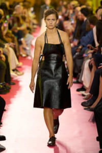 Model walking the runway in a black glossy halter-neck dress on a pink catwalk, audience blurred in the background.