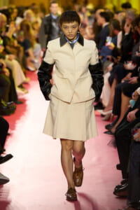 Model walks the runway in a cream suit with black leather sleeves and brown oxford shoes; pink catwalk with spectators.