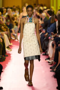 Model walks a pink runway in a cream beaded sleeveless dress with a colorful patterned scarf at the neck and brown oxford shoes, audience blurred in background.