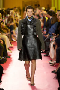 Model walking a fashion runway in a black leather A-line dress with a large ruffled bib over a blue shirt and olive-green sleeves, pink catwalk background.