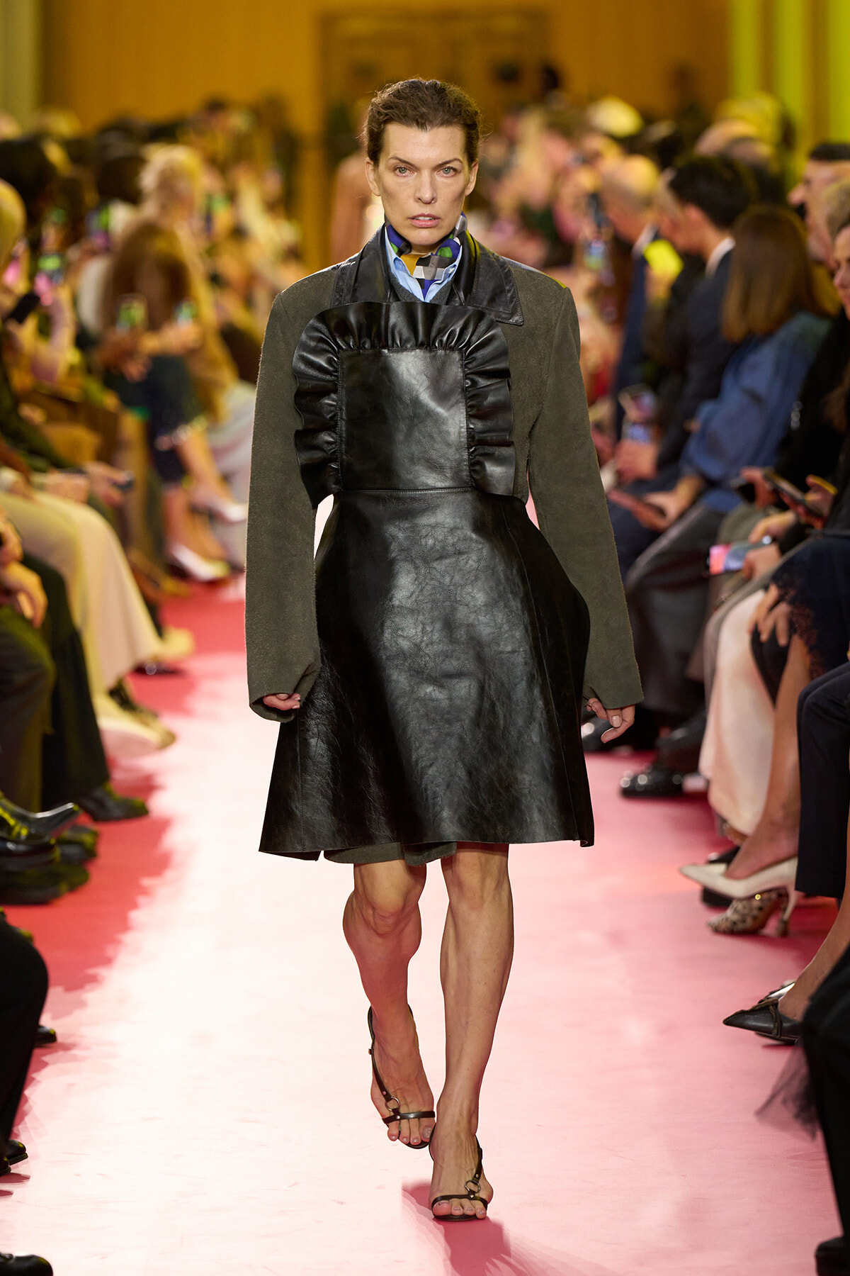 Model walking a fashion runway in a black leather A-line dress with a large ruffled bib over a blue shirt and olive-green sleeves, pink catwalk background.
