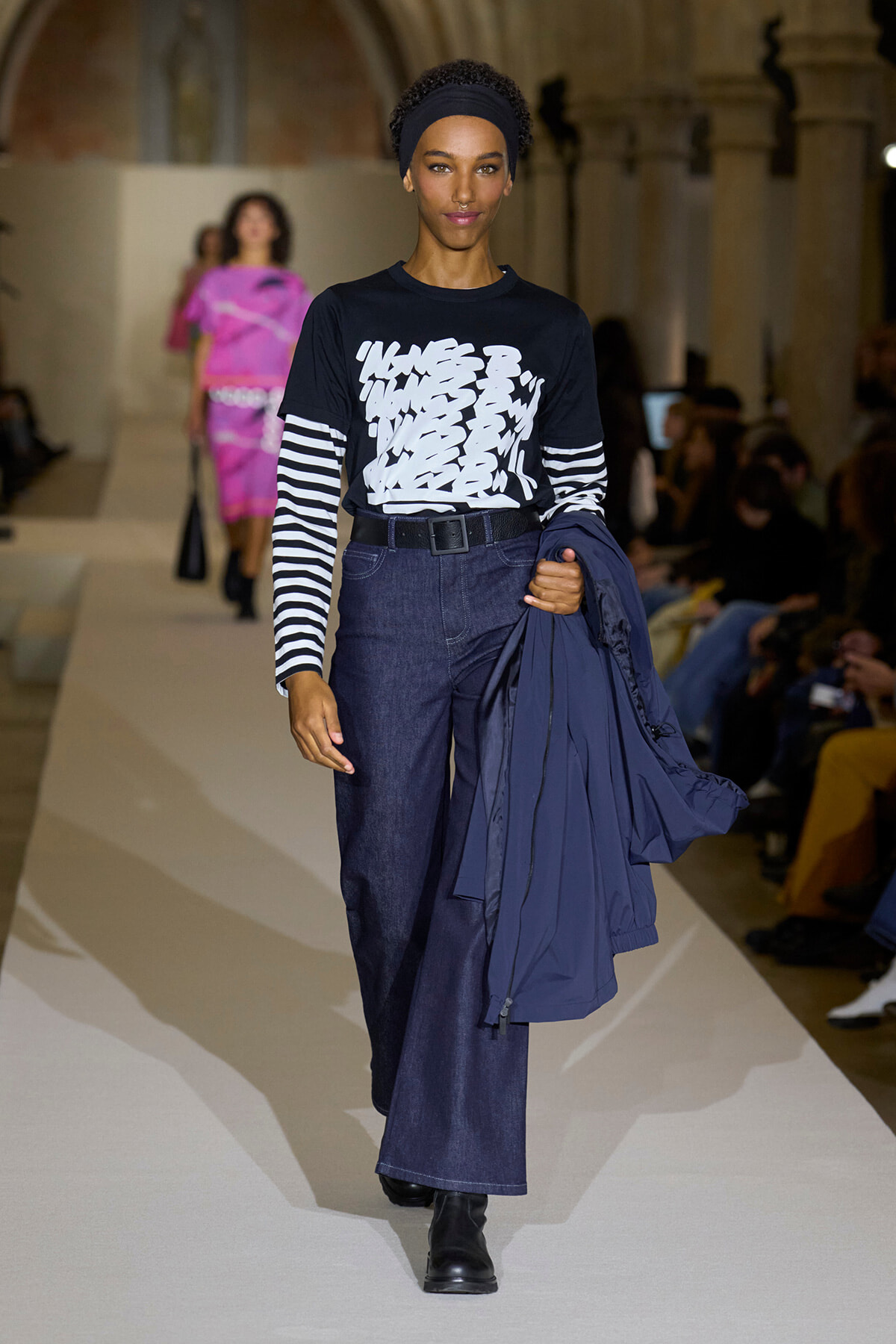 Model walking a runway in a black graphic tee with striped sleeves, high-waisted dark denim jeans, black belt, and black boots, holding a navy jacket over her arm.