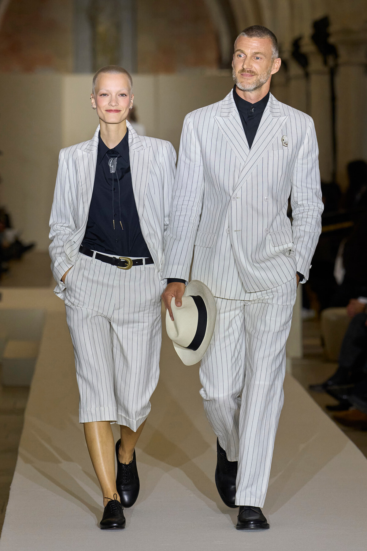 Two models walk a runway in matching light-gray pinstripe suits with navy shirts; the man holds a white fedora on his side.3a8# Wait, no emoji in ALT text.