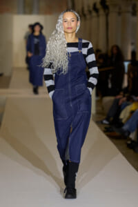 Model on a runway wearing a dark denim jumpsuit with a front zipper and large pockets, plus a black-and-gray striped top and long silver braided hair.