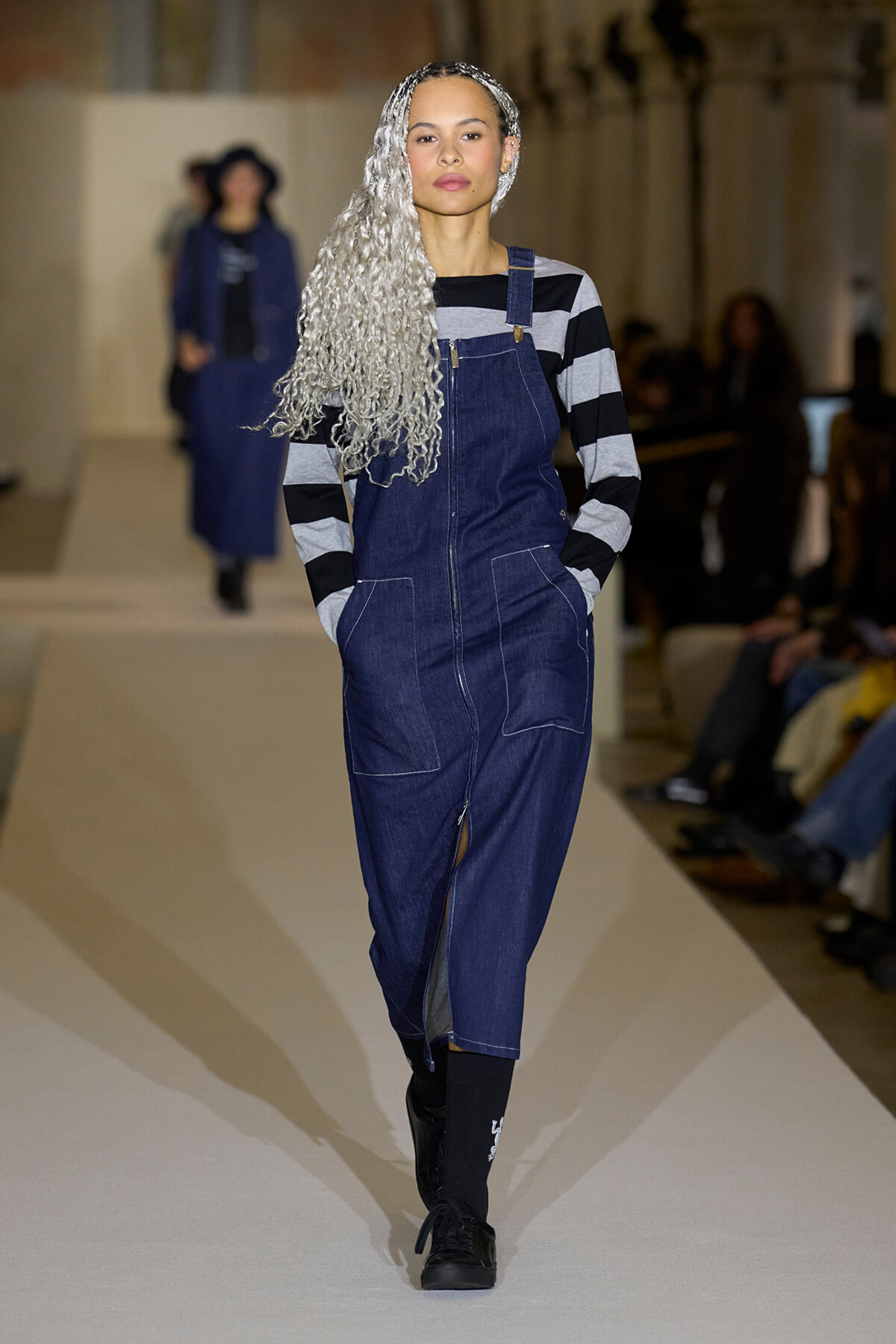 Model on a runway wearing a dark denim jumpsuit with a front zipper and large pockets, plus a black-and-gray striped top and long silver braided hair.