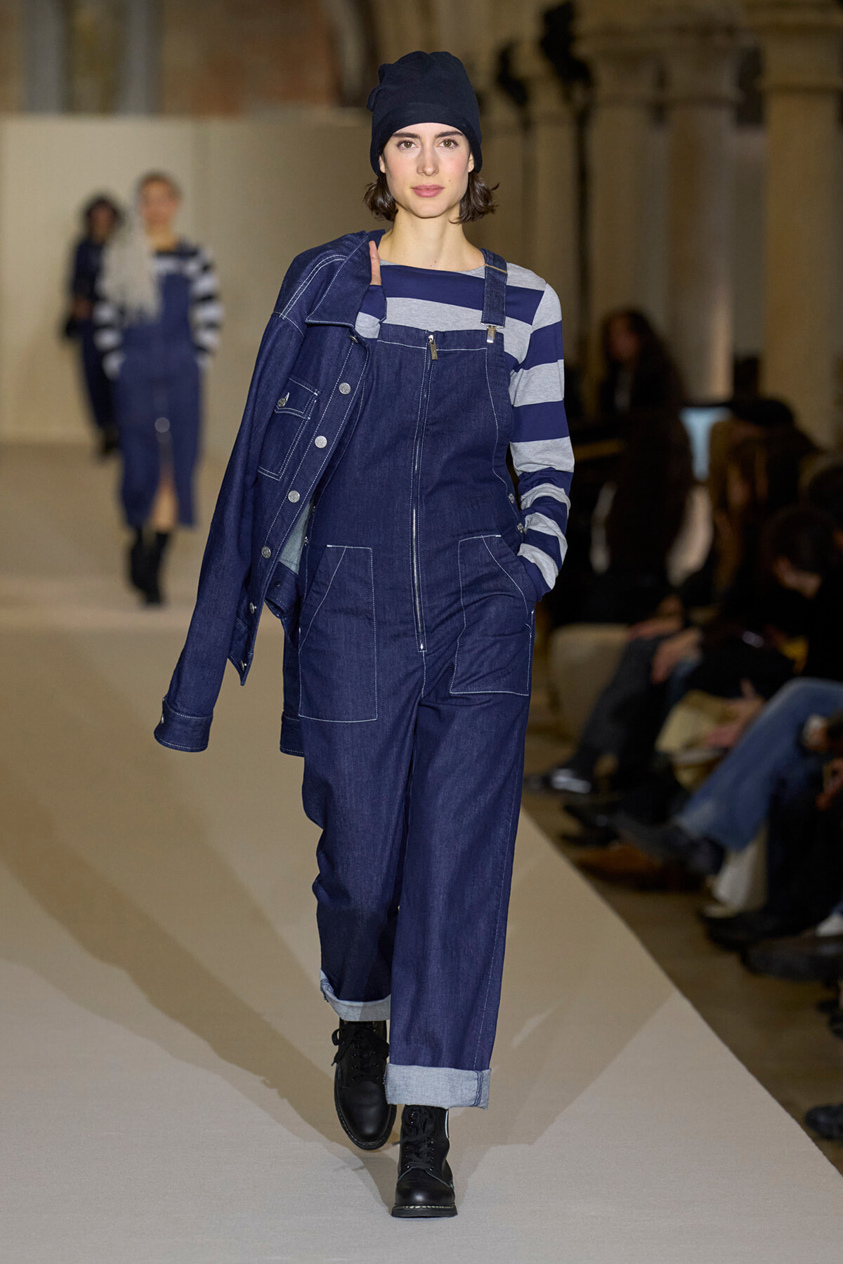 Model walking a fashion runway in dark denim overalls layered with a striped long-sleeve top and a dark beanie.