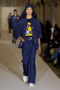 Model walks a fashion show runway in a denim-on-denim ensemble with a yellow graphic tee, cap, and white sneakers