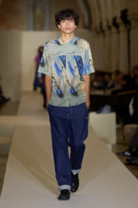 Male model walking a runway in a light abstract-print short-sleeve shirt and loose dark blue jeans with rolled cuffs, black shoes.