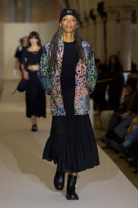 Model walks a fashion show runway in a multicolor circular-patterned jacket, black top and long black skirt, black boots, and a black beanie.