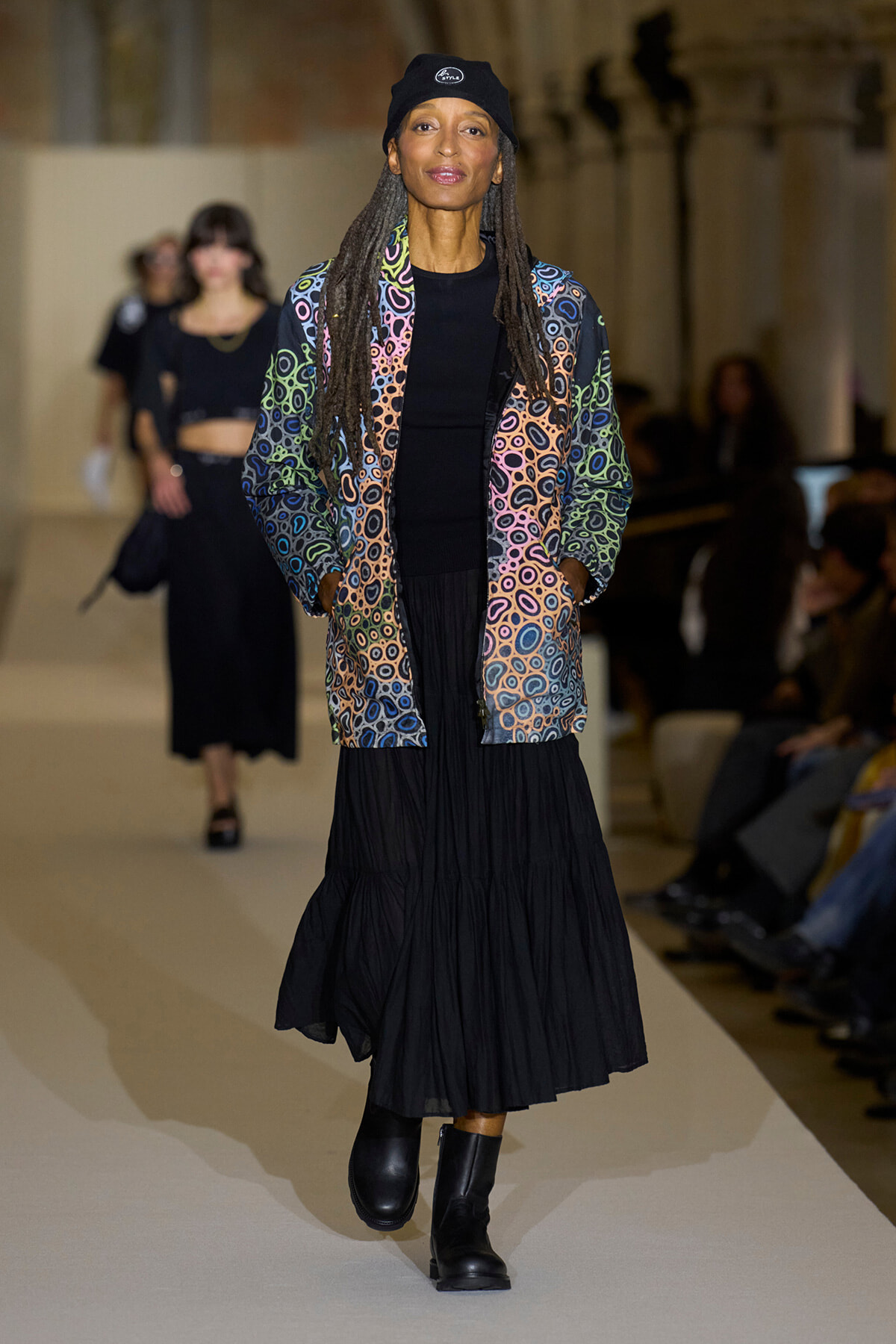 Model walks a fashion show runway in a multicolor circular-patterned jacket, black top and long black skirt, black boots, and a black beanie.